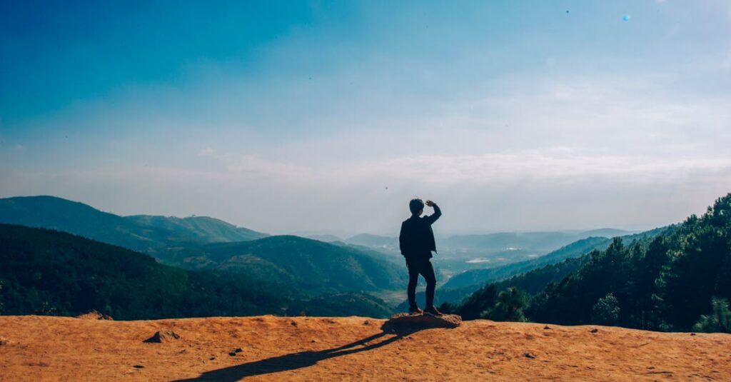 A lone traveler stands on a hilltop, gazing over a vast mountain landscape under a clear blue sky.